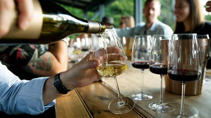 White wine being poured from a bottle into crystal glasses on a wooden table with guests in the background. White wine being poured from a bottle into crystal glasses on a wooden table with guests in the background.