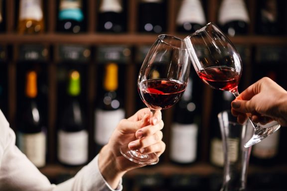 Wine Cellar Tasting Toast Two guests clinking glasses of red wine in front of a wine cellar rack background.